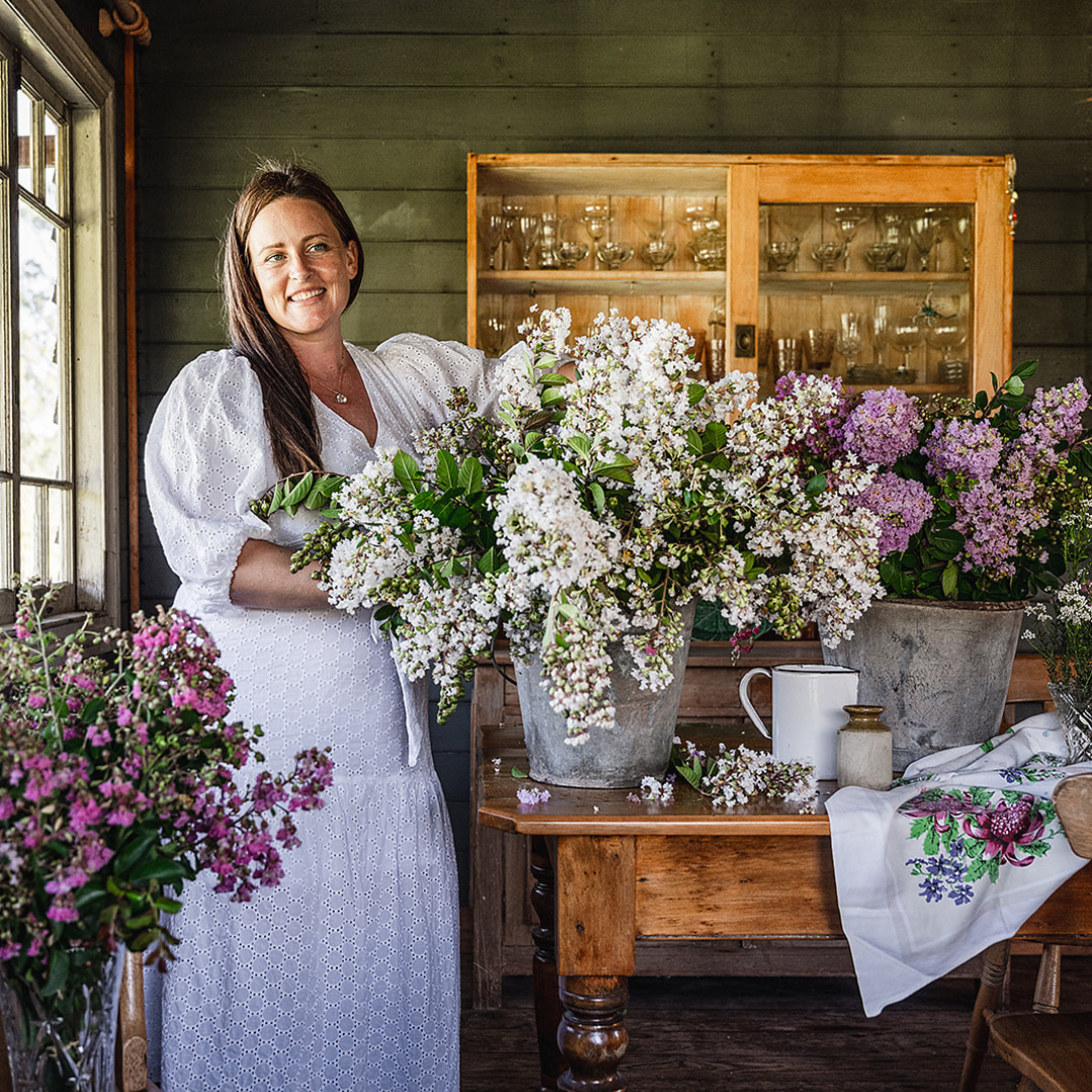 A woman in a white dress stands indoors next to a wooden table filled with large bouquets of flowers in metal buckets. The room has a rustic feel with wooden furniture and a cabinet in the background.